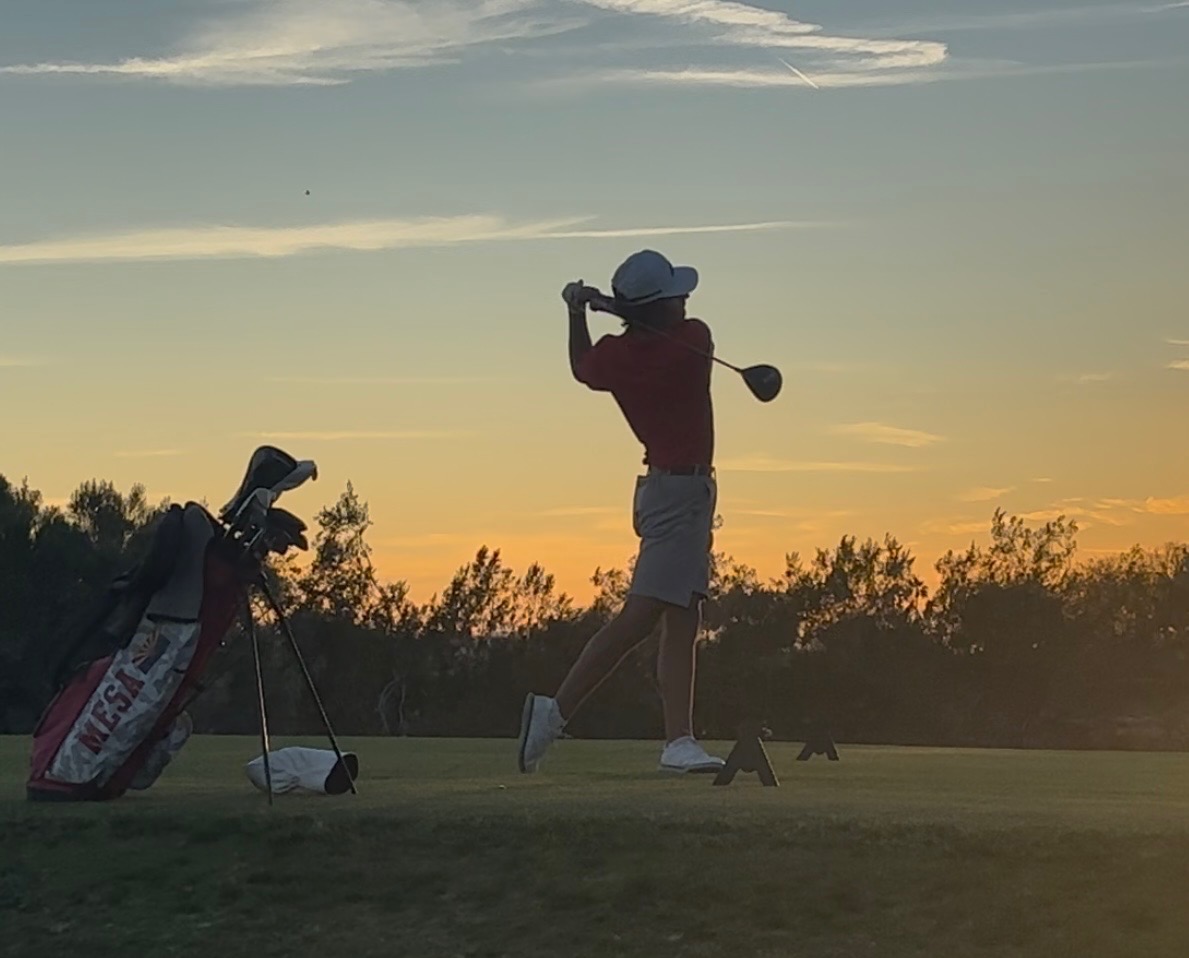 A golfer follows through on a tee shot during sunset, silhouetted against the evening sky. A golf bag with “Mesa” visible on the side stands nearby as trees line the background of the course.
