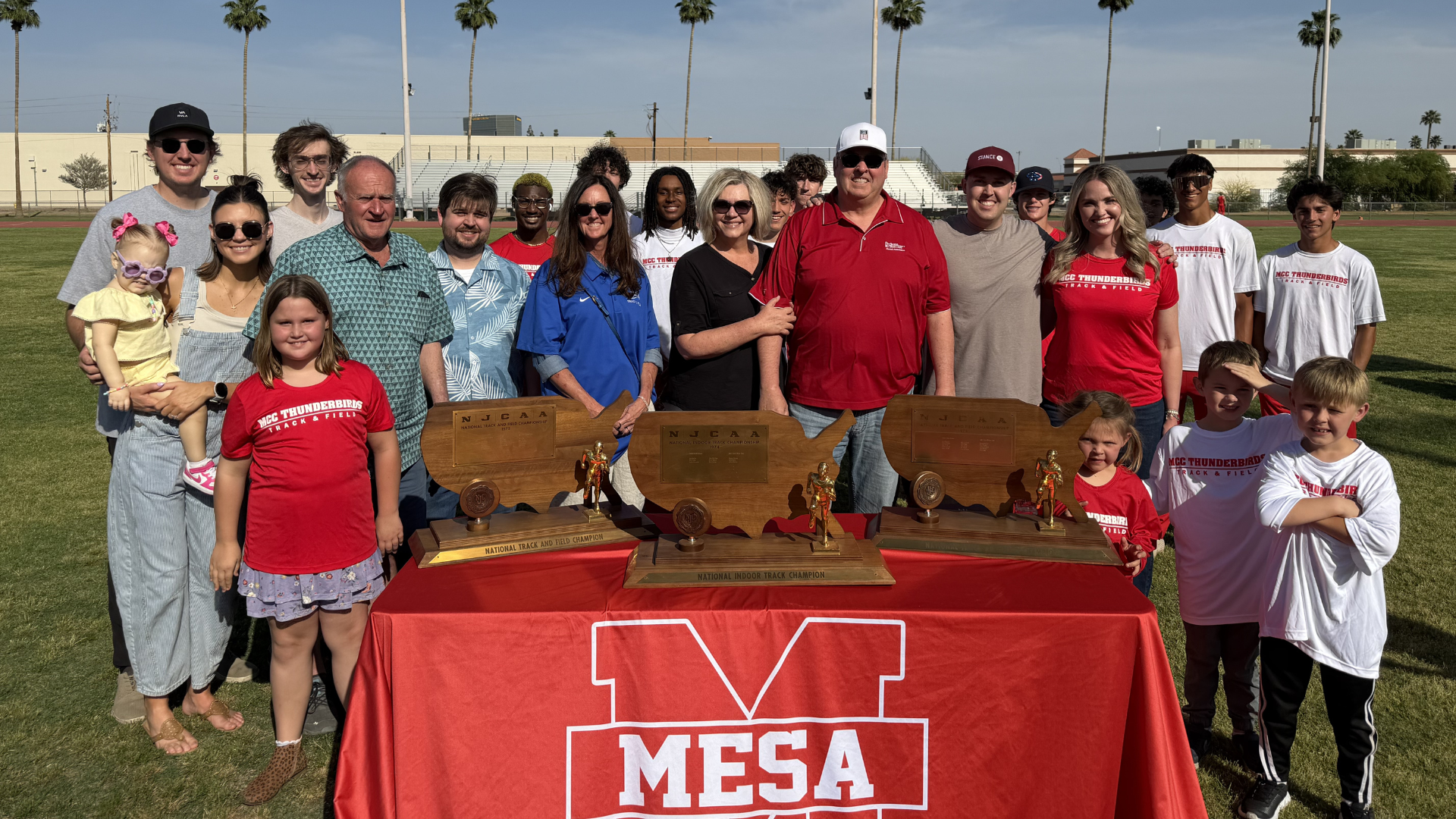 The Jackson family stands together on the infield at Mesa Community College, surrounded by MCC Track & Field athletes and staff, posing behind a table displaying multiple NJCAA national championship trophies in honor of Carvel Jackson.
