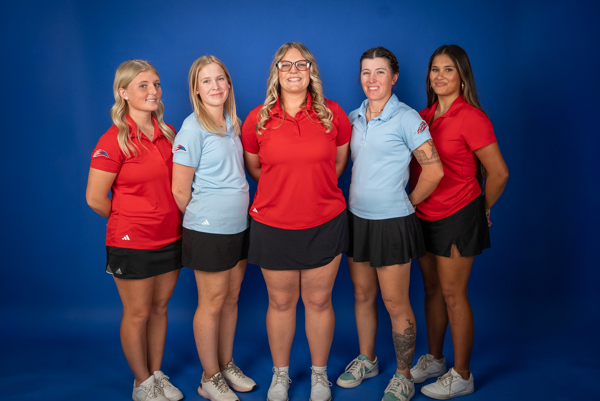 Five members of the Mesa Community College women’s golf team stand together for a posed team photo against a blue backdrop. The athletes wear Mesa polos in red and light blue with black skirts and white golf shoes. Three athletes wear red polos while two wear light blue polos. The group smiles toward the camera with hands behind their backs during media day portraits.
