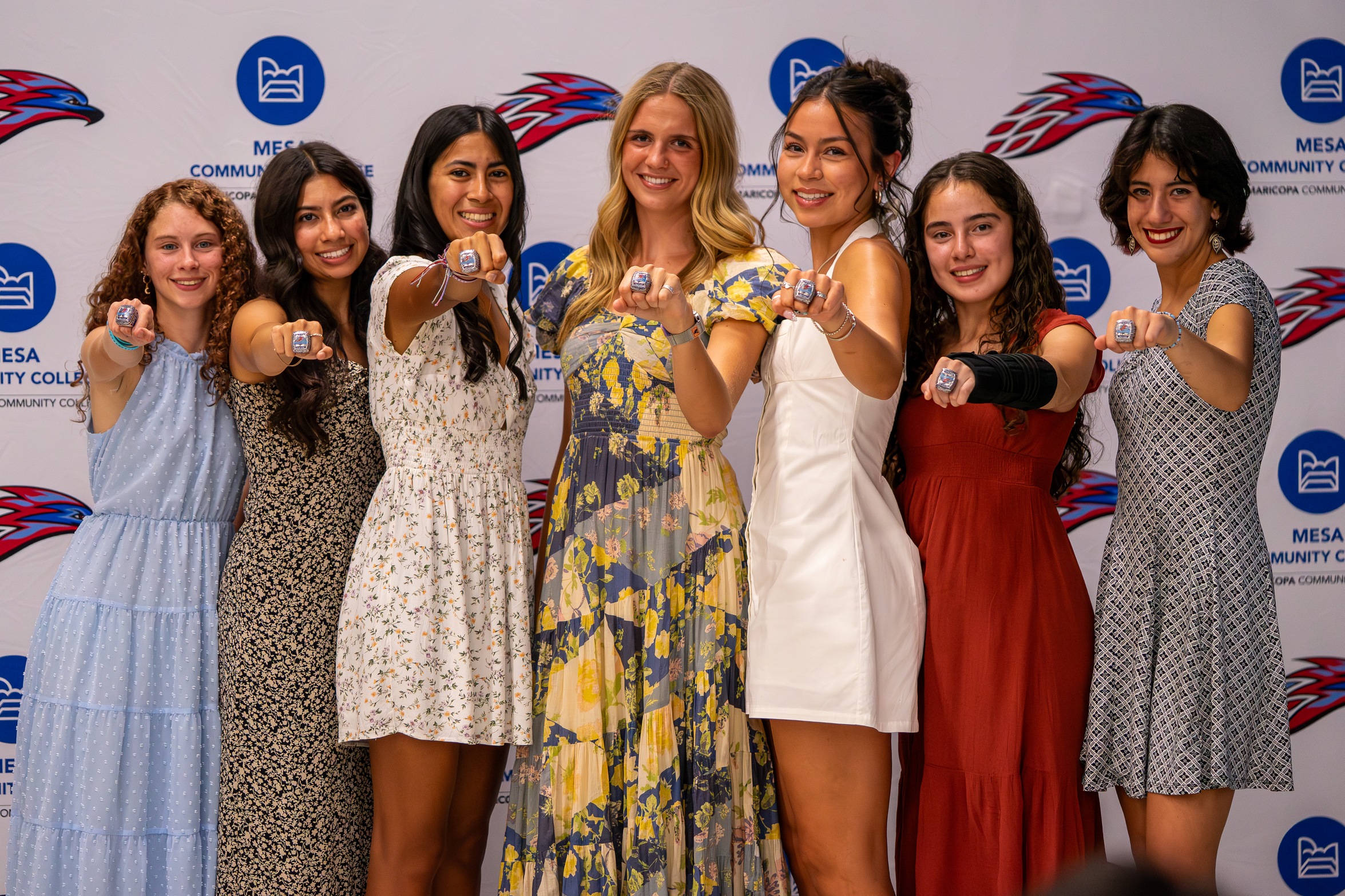 A photo of 7 girls holding their arms up with rings on their fingers all smiling with white background contacting Mesa CC logo & Thunderbird logo.